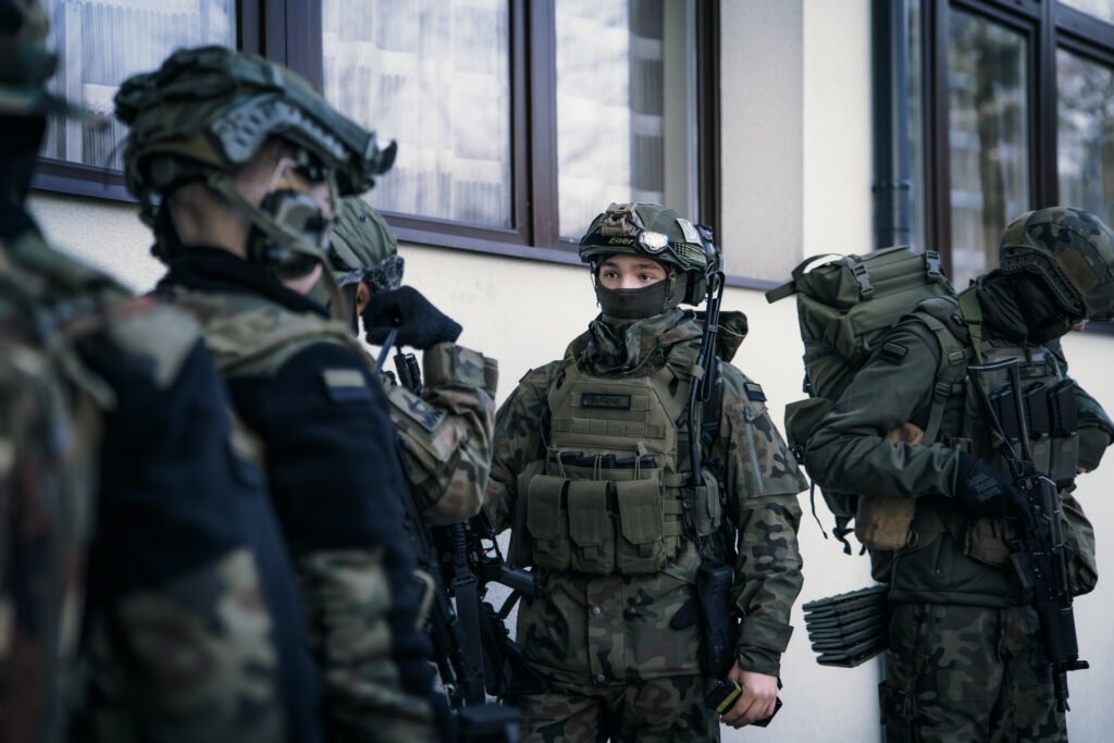 Grupa uzbrojonych Strzelców w kamizelkach typu plate-carrier i hełmach balistycznych stoi na tle budynku.