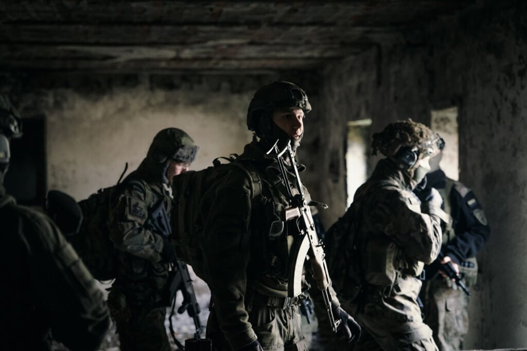 Grupa uzbrojonych strzelców, w hełmach i kamizelkach typu plate-carrier, z plecakami patrolowymi na plecach, stoi w opuszczonym budynku.