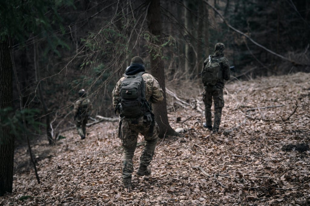 Widok z tyłu na grupę uzbrojonych Strzelców idących jeden za drugim przez zarośnięte lasem zbocze. Podłoże pokrywają uschnięte liście.