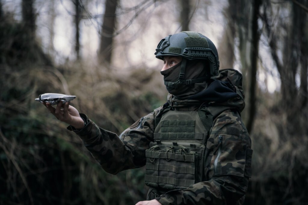 Strzelec ubrany w kurtkę w kamuflażu wz. 93, kamizelkę typu plate-carrier oraz hełm balistyczny trzyma w prawej ręce gotowy to startu dron typu quadcopter.